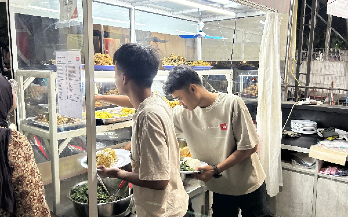 Foto Dua orang karyawan RM Padang Suku Jambak tengah meracik porsi nasi beserta lauk pauk pesanan pelanggan di balik etalase kaca pada malam hari, guna memastikan sajian disiapkan dengan cepat di tengah tingginya minat pembeli. (Fabio Pratama/Ketik.com)