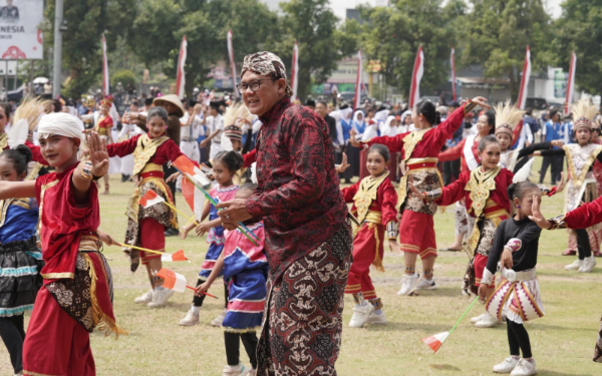 Foto Wabup Sleman Danang Maharsa (tengah), mengenakan busana adat Jawa dan blangkon, tersenyum lebar sambil memegang bendera Merah Putih kecil. Di sekelilingnya, penari-penari muda dengan kostum tari tradisional merah mencolok ikut menari dalam semangat kemerdekaan. (Foto: Pemkab Sleman for Ketik.com)